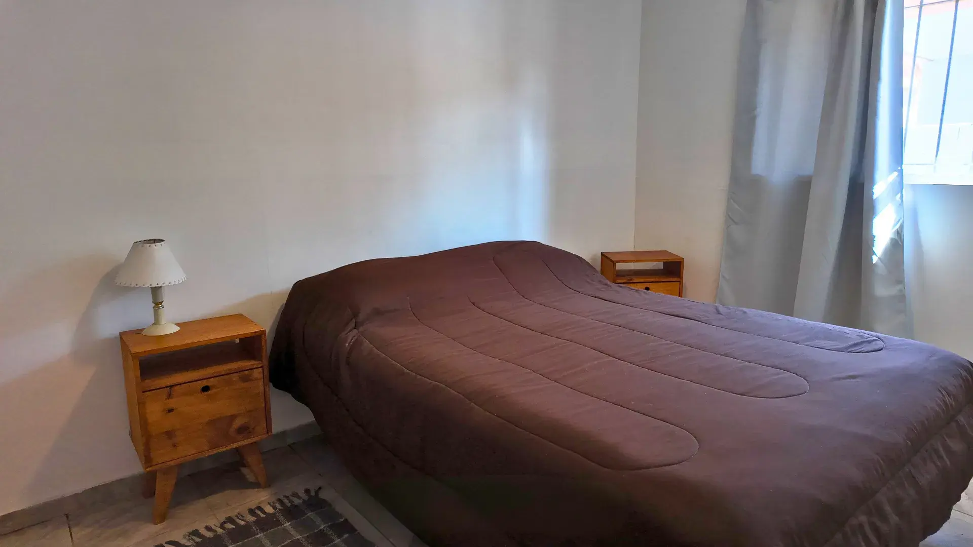 Dormitorio con cama matrimonial, dos mesas de luz de madera y cortinas en departamento en alquiler en Villa del Dique